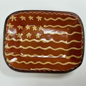 May include: A rectangular ceramic tray with a glossy, dark brown glaze featuring a crackle finish. The surface displays a stylized design of a flag with cream-colored wavy horizontal stripes and a cluster of small, five-pointed stars in the corner.