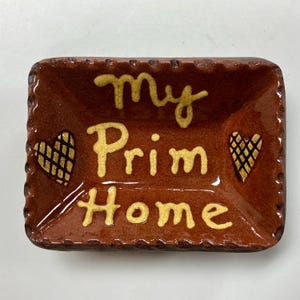 May include: A rectangular, brown ceramic dish with the words "My Prim Home" in yellow script. Two yellow hearts with black crosshatch patterns are on either side of the text. The dish has a scalloped edge.