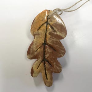 May include: Ceramic leaf pendant with a naturalistic design. The pendant features a brown and tan color scheme with black vein detailing. A twine loop is attached for hanging. This decorative item is suitable for fall-themed decor.