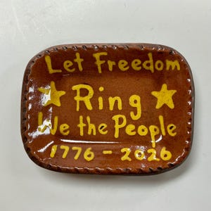 May include: A rectangular, brown ceramic dish with the words "Let Freedom Ring We the People 1776-2020" in yellow. The dish has a stitched border and two yellow stars.