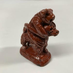 May include: A small, hand-crafted ceramic sculpture of two animals, one on top of the other. The sculpture is a reddish-brown color and has a textured surface.