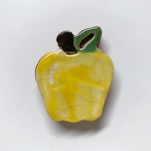 May include: A yellow ceramic apple with a green leaf and a brown stem. The apple has a textured surface.