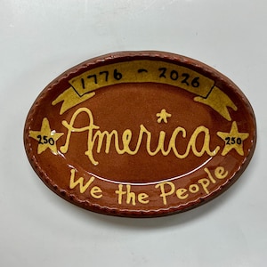 May include: Oval-shaped, brown ceramic plate with a decorative design. The plate features the word "America" in yellow script, along with the text "We the People." It also includes the years "1776 - 2026" and two stars with the number "250."