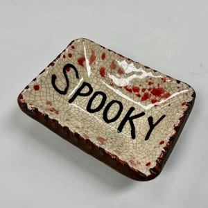 May include: A rectangular ceramic dish with a cream-colored crackle glaze finish, featuring hand-painted red splatters and the word SPOOKY written in bold black lettering across the center. The edges of the dish are finished in a dark brown tone.