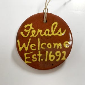 May include: A round, brown ceramic ornament with the words "Ferals Welcome Est. 1692" in yellow script. The ornament has a string for hanging.