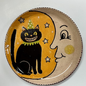 May include: A round ceramic plate with a crackle glaze finish featuring a black cat wearing a green ruffled collar and a green polka-dot party hat. The cat sits against a large yellow moon shape with white stars, next to a crescent moon face.