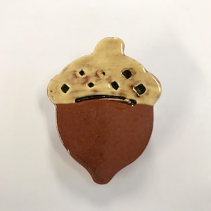 May include: A ceramic acorn figurine with a light tan top and a brown bottom. The top has small, dark square cutouts and a black line separating the top and bottom. The acorn is a decorative item.