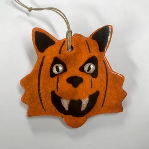 May include: Orange ceramic Halloween ornament shaped like a jack-o'-lantern with black accents. The ornament has a string for hanging and features a cracked glaze finish. The jack-o'-lantern has black eyes, nose, and mouth with white teeth.