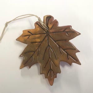 May include: A decorative maple leaf ornament with a brown and gold finish. The leaf has detailed veining and a twine loop for hanging. The ornament is likely made of ceramic or a similar material.