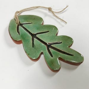 May include: A ceramic leaf ornament with a green glaze and black vein details. The leaf has a brown edge and is attached to a natural twine string. The ornament is a decorative item.