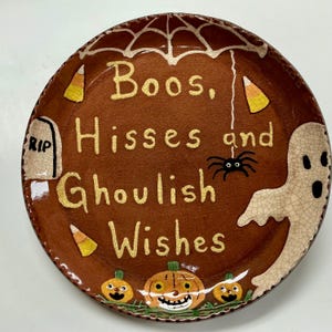 May include: A round ceramic plate with a brown crackle glaze featuring the text Boos, Hisses and Ghoulish Wishes in yellow. The design includes a white spider web, a black spider, a tombstone with RIP, a white ghost, candy corn, and pumpkins.