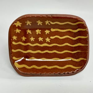 May include: A rectangular ceramic dish with rounded corners, featuring a glossy brown glaze and a crackle finish. The surface is decorated with a stylized yellow pattern of wavy horizontal lines and small stars in the upper left corner.