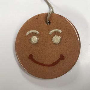 May include: A round, brown ceramic ornament with a gingerbread man face. The ornament has white eyes, white eyebrows, and a red smiling mouth. A string is attached at the top for hanging.