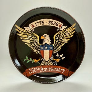 May include: A round, dark brown decorative plate with a painted design. The design features a bald eagle with outstretched wings, a shield, and the text "1776-2026" and "250th Anniversary". The eagle is in shades of yellow, white, and brown.