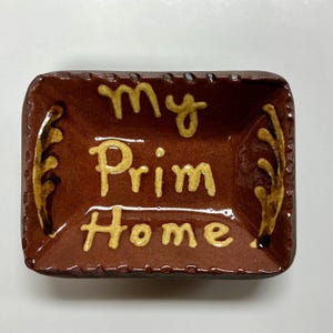 May include: A rectangular, brown ceramic dish with the words "my Prim Home" in yellow script. The dish has a slightly raised rim with decorative accents and is likely used for decorative purposes.