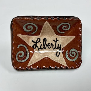 May include: A rectangular ceramic tray with a glossy brown finish and a crackle glaze texture. The center features a large, cream-colored star with the word Liberty written in black script. Four light blue spiral designs accent the corners.