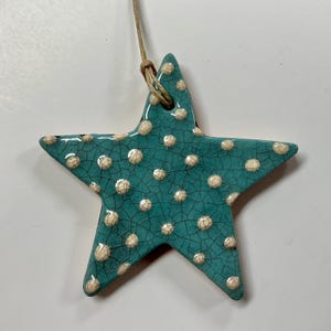 May include: A turquoise ceramic star-shaped ornament with a crackle glaze and white polka dots. The star is suspended by a natural twine loop, suitable for hanging. This decorative item is ideal for nautical or coastal themed decor.