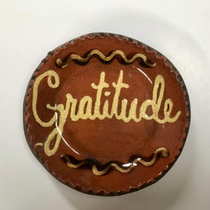 May include: A small, round, ceramic plate with a brown base and a cream-colored inscription of the word "gratitude". The plate has a decorative wavy border and a textured surface. A unique piece for home decor.