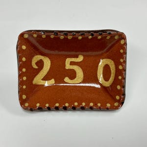 May include: A rectangular ceramic dish with a glossy dark brown glaze and a visible crackle finish. The center features the number 250 in a light cream color, surrounded by a decorative border of small cream-colored dots along the edges.