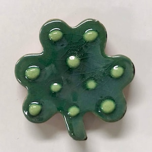 May include: A ceramic shamrock with a glossy, crackled green finish. The shamrock features raised, light green dots and a small stem. The design is a stylized representation of a four-leaf clover.