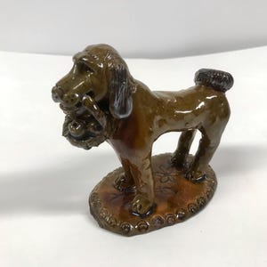 May include: A brown ceramic dog figurine with a brown and white tail. The dog is holding a brown and white basket with a brown and white bird in its mouth. The dog is standing on a brown ceramic base with a brown and white design.
