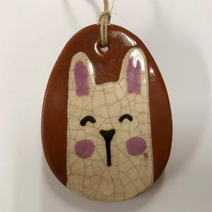 May include: A ceramic Easter egg ornament with a brown background and a white bunny face with pink ears, nose, and cheeks. The bunny has a black smile and nose.