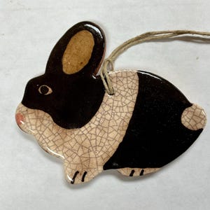 May include: A ceramic bunny ornament with a black and cream color scheme. The bunny has a black head and back, with cream-colored body and ears. A small pink nose and black feet complete the design. A twine loop is attached for hanging.