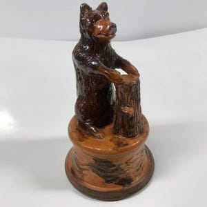 May include: A ceramic bear figurine standing on a tree stump. The bear is brown and has a textured surface. The tree stump is also brown and has a textured surface.