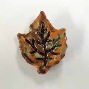 May include: A ceramic leaf-shaped decorative item. The leaf features a detailed black vein design against a mottled background of orange, brown, and green. The item has a glossy finish and is likely handmade.