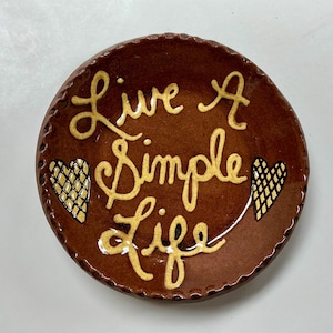 May include: A round, brown ceramic dish with the words "Live A Simple Life" in yellow script. Two yellow hearts with black crosshatch patterns are on either side of the text. The dish has a textured rim.