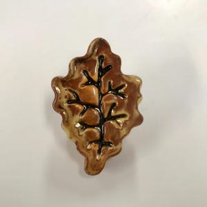 May include: A ceramic leaf-shaped decorative item with a brown and tan glaze. The leaf features a detailed black tree branch design in the center. The item is likely a decorative accent for home decor.