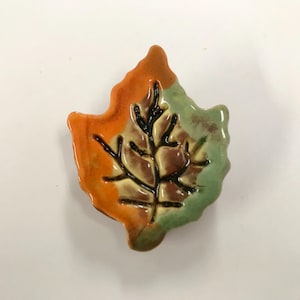 May include: A ceramic leaf-shaped decorative item. The leaf is divided into three sections: orange, green, and brown. Black lines depict the veins of the leaf. The item is likely a fall-themed decoration.