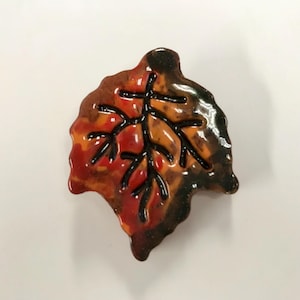 May include: A ceramic leaf-shaped pin with a glossy finish. The pin features a color palette of red, orange, and brown, with black veins. The leaf is a fall-themed decorative item.