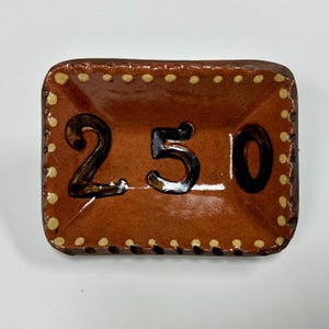 May include: A rectangular brown ceramic tray with a glossy finish, featuring the black painted number 250 in the center. The edges are decorated with a border of small, cream-colored dots, and the surface has a subtle, fine crackle texture.