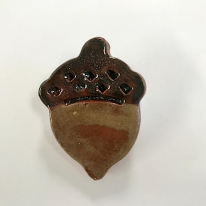 May include: A ceramic acorn figurine with a glossy, dark brown cap and a lighter brown body. The cap has diamond-shaped indentations. The figurine is set against a white background.