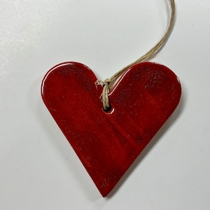 May include: A glossy, heart-shaped ornament in a vibrant red hue. The wooden heart has a small hole at the top, threaded with a natural twine for hanging. The ornament is a simple, yet elegant decorative piece.
