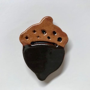 May include: A ceramic acorn ornament with a brown glaze on the bottom half and a light brown glaze on the top half. The top half has a textured surface and a few small holes.