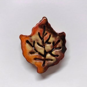 May include: A ceramic leaf-shaped decorative item with a glossy finish. The leaf features a blend of orange, brown, and cream colors, with dark brown veins. This fall-themed decor piece is likely a brooch or a decorative accent.