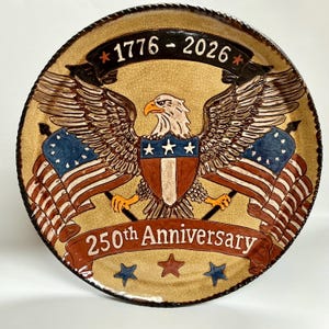 May include: A round ceramic plate with a crackled tan glaze featuring a central bald eagle with spread wings, a shield, and two American flags. A black banner at the top reads 1776-2026, and a red banner at the bottom reads 250th Anniversary.