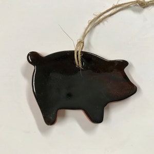 May include: A ceramic pig ornament with a glossy dark brown finish. The pig has a simple, stylized design with visible legs and a curled tail. A natural twine loop is attached for hanging.