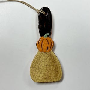May include: A ceramic hanging ornament shaped like a broom with a black handle, an orange pumpkin accent, and a cream-colored base. The entire piece features a distinct crackle glaze finish and includes a thin twine string attached at the top.