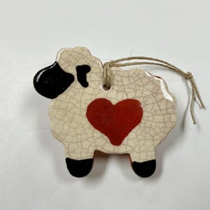May include: A ceramic sheep ornament with a cream-colored body and a red heart. The sheep has black accents on its head and feet, and a jute string for hanging. The ornament has a crackle glaze finish.
