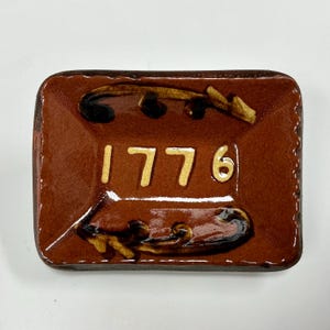 May include: A rectangular brown ceramic dish with a glossy finish. The center features the cream-colored text 1776, flanked by dark brown, hand-painted scrollwork designs above and below the numbers. The edges of the dish are slightly raised.