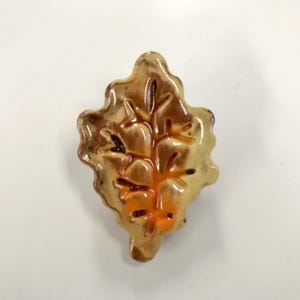 May include: A ceramic leaf-shaped decorative item. The leaf is primarily a warm, golden-brown color with darker brown veins and accents. The base of the leaf features an orange hue. The item has a glossy finish and is set against a white background.