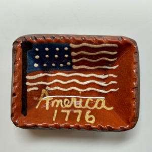 May include: A rectangular ceramic tray with a glossy, crackled brown glaze. The center features a painted American flag with a dark blue canton, white stars, and wavy white stripes. The text "America 1776" is written in cream-colored script.