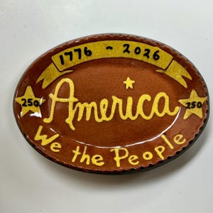 May include: An oval ceramic dish with a brown crackle glaze featuring yellow accents. It displays the text 1776 - 2026 on a banner, the word America, and We the People. Two yellow stars on the sides are marked with the number 250, and one small star.