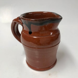 May include: A brown ceramic pitcher with a black glaze around the rim. The pitcher has a rounded body and a handle.