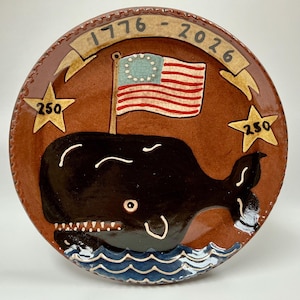 May include: A round ceramic plate with a brown glaze featuring a black whale swimming in blue waves. Above the whale is an American flag with thirteen stars and the text 1776 - 2026 on a banner. Two yellow stars with the number 250 are on the sides.