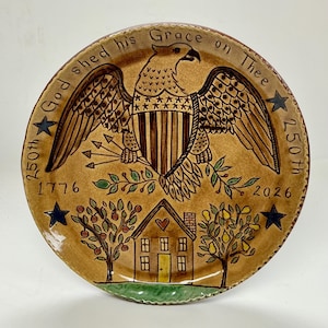 May include: A decorative plate with a patriotic design. The plate features a brown eagle with outstretched wings, a shield, and the text "God shed his Grace on Thee." The plate also includes a house, trees, stars, and the dates 1776 and 2026.