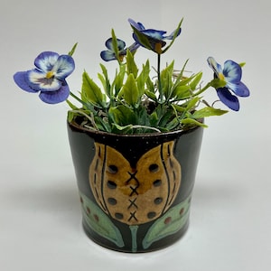 May include: A small, black ceramic flower pot with a decorative, stylized floral design in brown and green. The pot contains artificial blue and white pansies with green leaves, creating a vibrant display. The pot is approximately 4 inches tall.